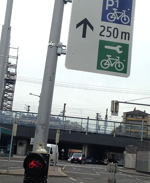 Schild am Fahrradampelmast mit Piktogrammen Radgarage und Radservice, Entfernungsangabe und Richtungspfeil