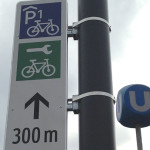 Schild am Laternenmast mit Piktogrammen Radgarage und Radservice, Entfernungsangabe und Richtungspfeil