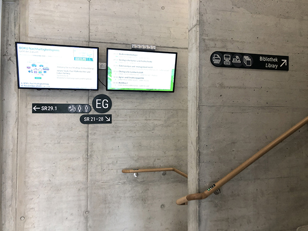 Dunkle Schilder mit weißer Schrift montiert auf Rohbetonwand unter zwei Bildschirmen